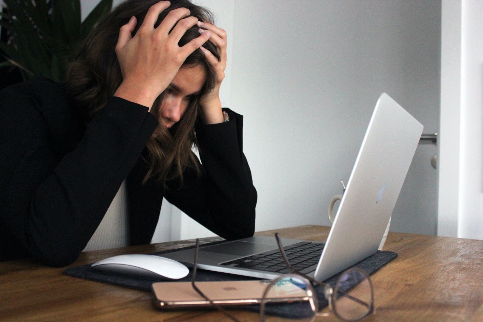 Writer’s block can be induced by perfectionism, distractions, fear, or stress. A woman in a black sweater sitting with her head in her hands, looking at her computer screen with a frustrated facial expression.