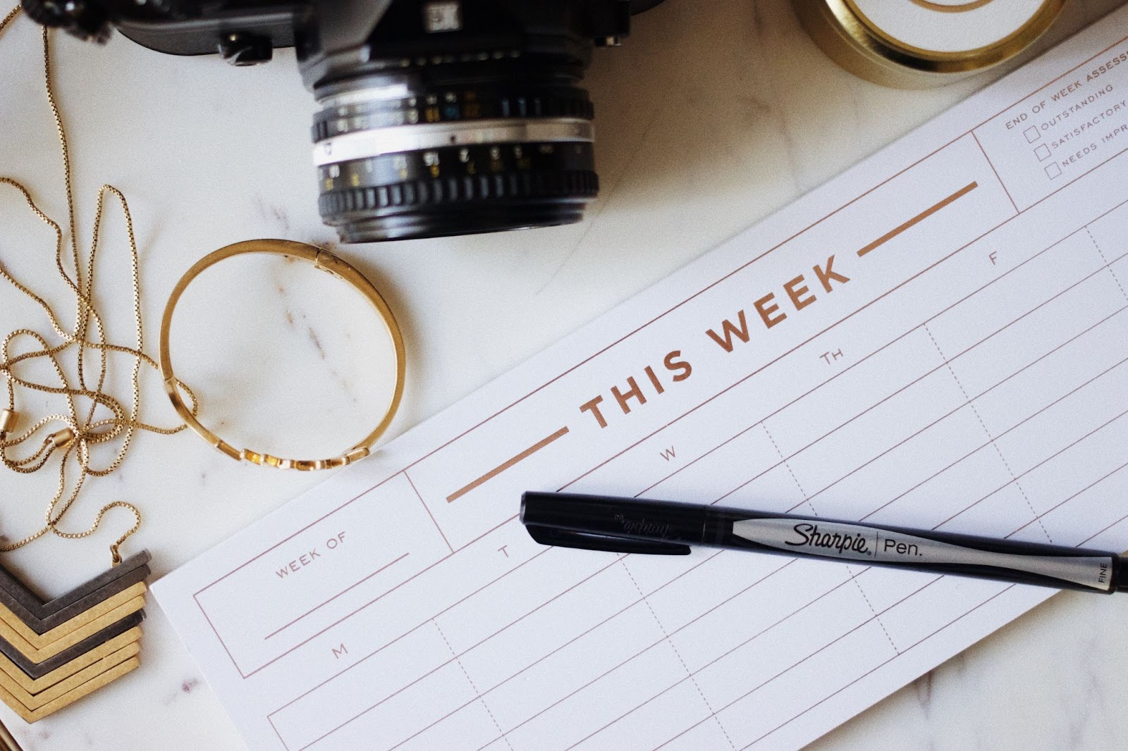 Setting aside time to write blog posts, either daily or weekly, will ensure that writing becomes a regular habit. A calendar on a white marble desk surrounded by a camera, jewelry, and a Sharpie pen.