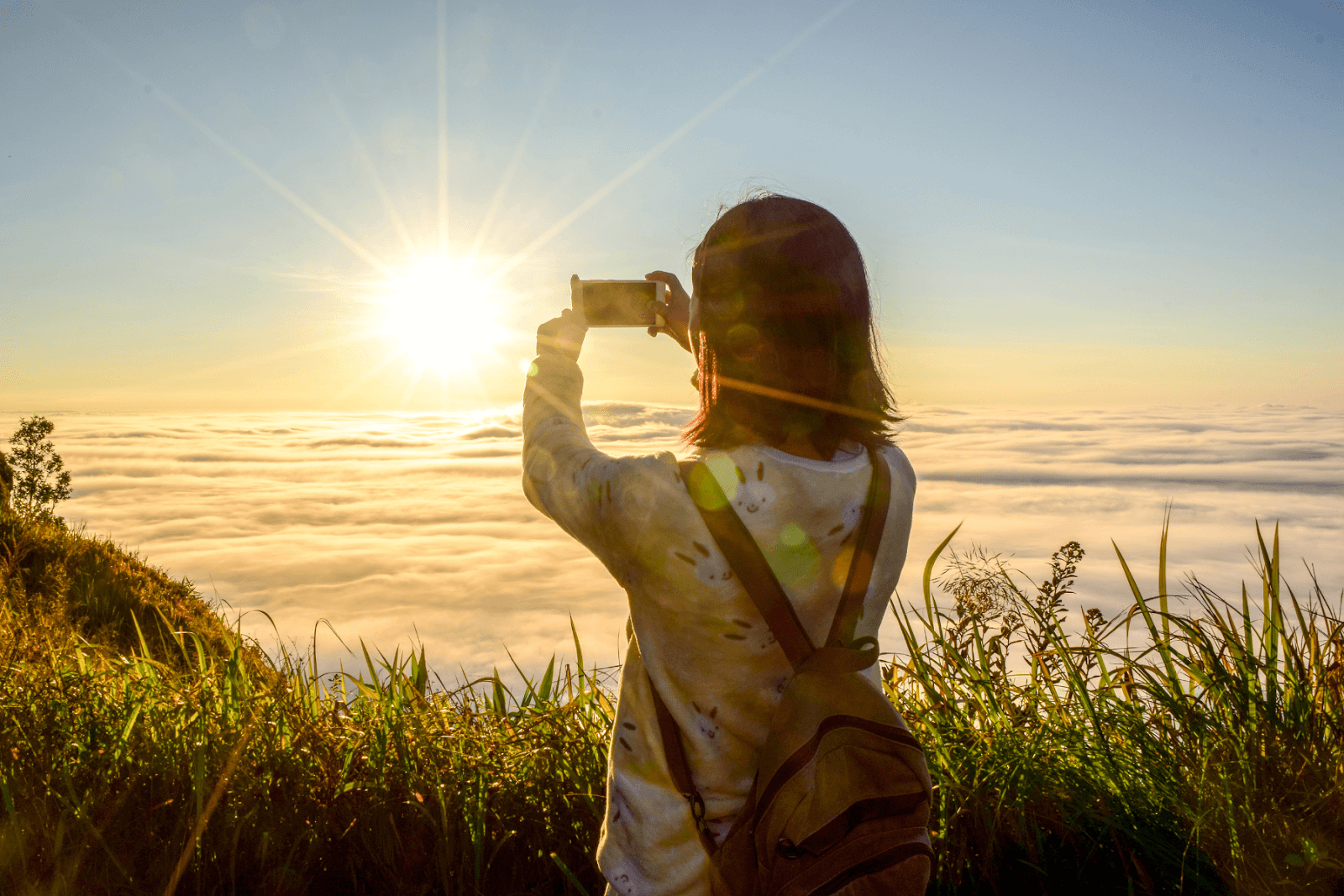 Visual search AI is revolutionizing how travelers discover their next destinations. The photo displays a woman taking a photo of a sunset high above the clouds. She is surrounded by green grass on a hilltop as the sun plunges beneath the scattered clouds.