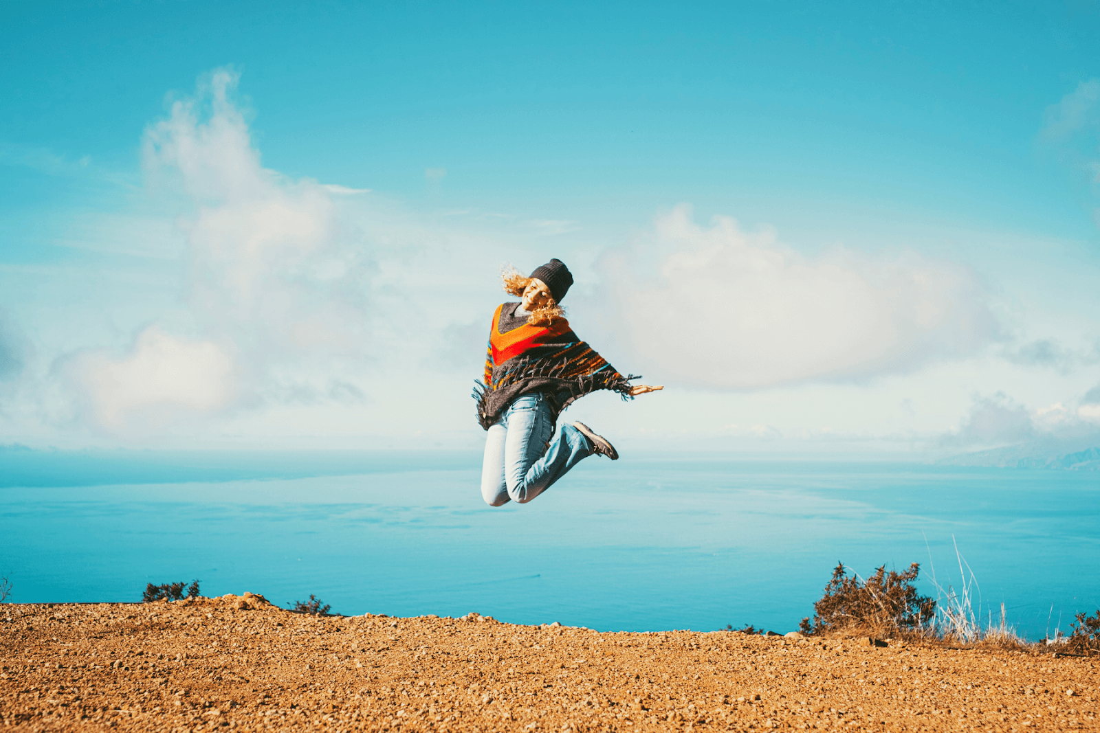 Travelers are discovering how beneficial visual search AI is in the planning process. The image displays a woman jumping into the air on top of a mountain with a beautiful ocean in the background. The mountaintop is dirt with a few shrubs, while the woman wears a beanie, jeans, and a poncho.