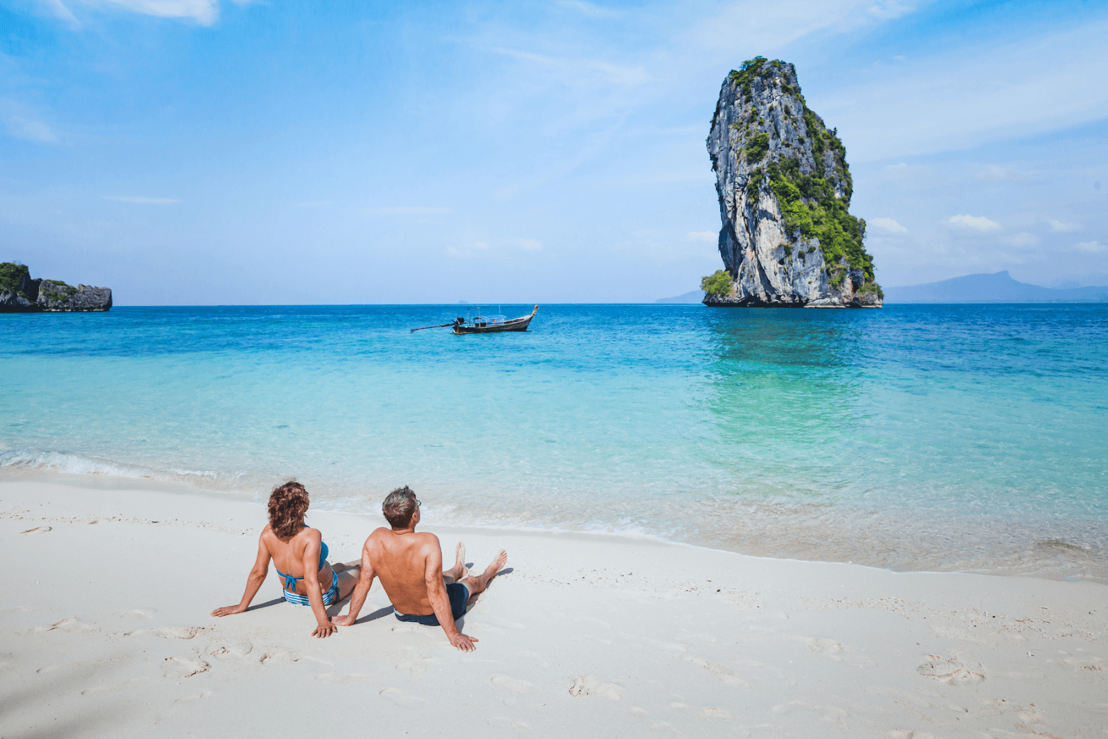 It’s easy to get inspired using visual search. The image displays a couple relaxing on a beach in Asia. The beach has white sand with turquoise waters, and a large rock jets upward from the ocean floor with a small boat nearby.