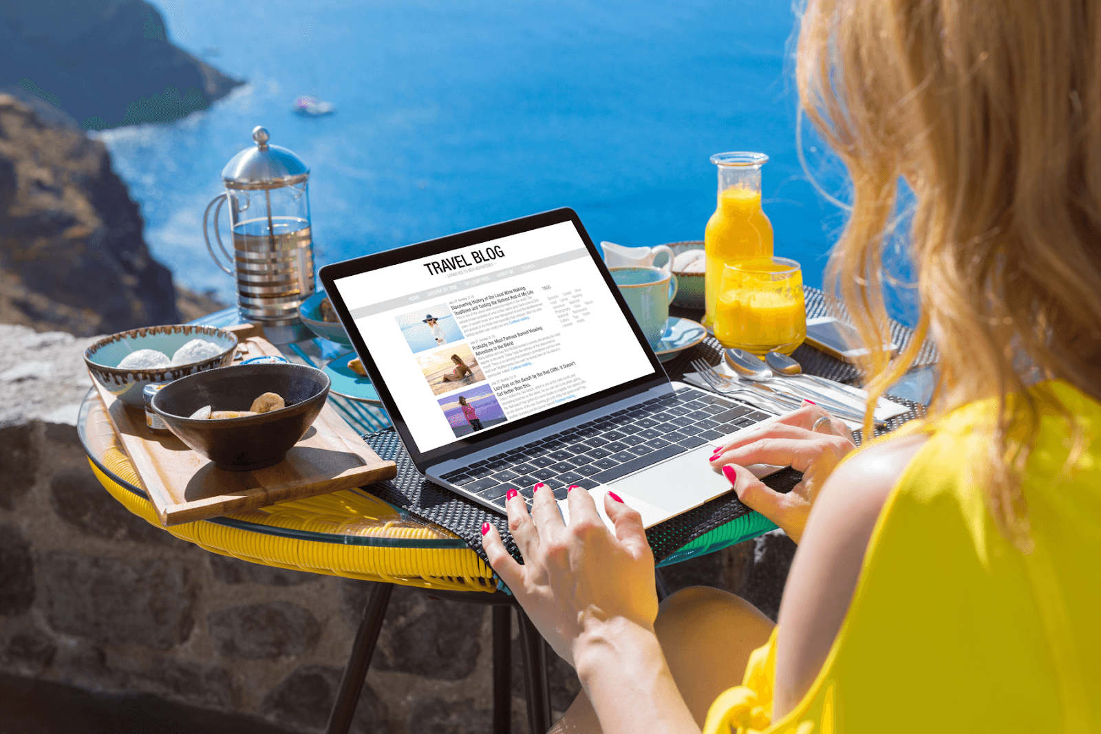 Travel bloggers are incorporating visual search into their blogging strategies. The photo displays a woman sitting at a small table with food, drinks, and a laptop. She is working on her travel blog as she overlooks a beautiful cliffside before plunging below into the ocean. A catamaran is in the distance floating on the water.